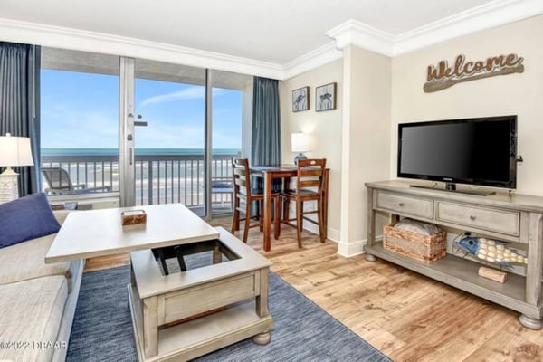 Beachfront condo living room with ocean view balcony, TV, dining table, and welcome sign